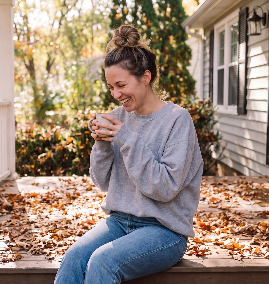 A caucasian woman in her mid-30's wearing a grey sweatshirt and jeans, sitting on the porch stair with a hot beverage, enjoying the fall weather.
