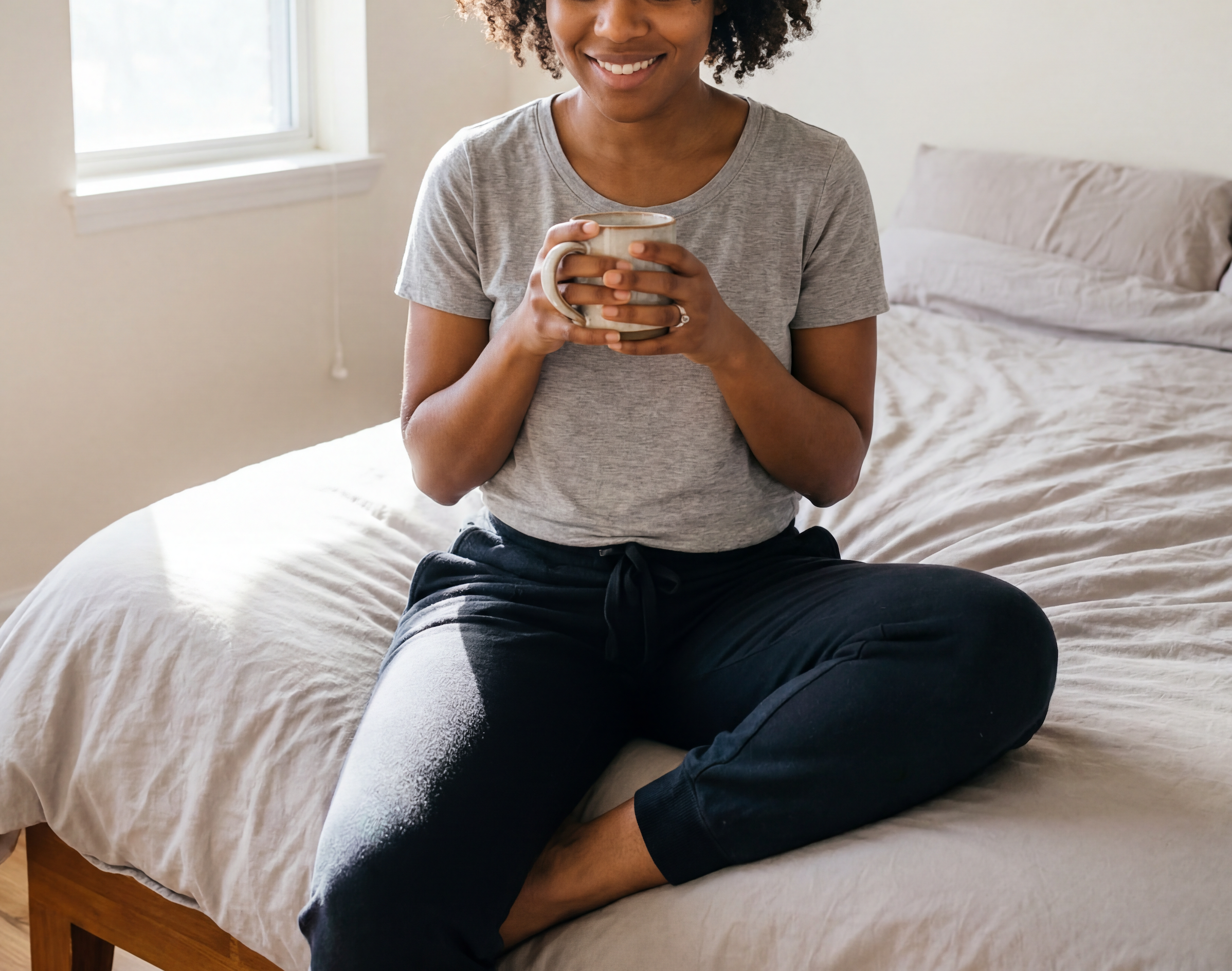 A black woman in her early 30's wearing a grey t-shirt and navy blue pants sitting on her bed, holding a mug. She looks rested and refreshed.