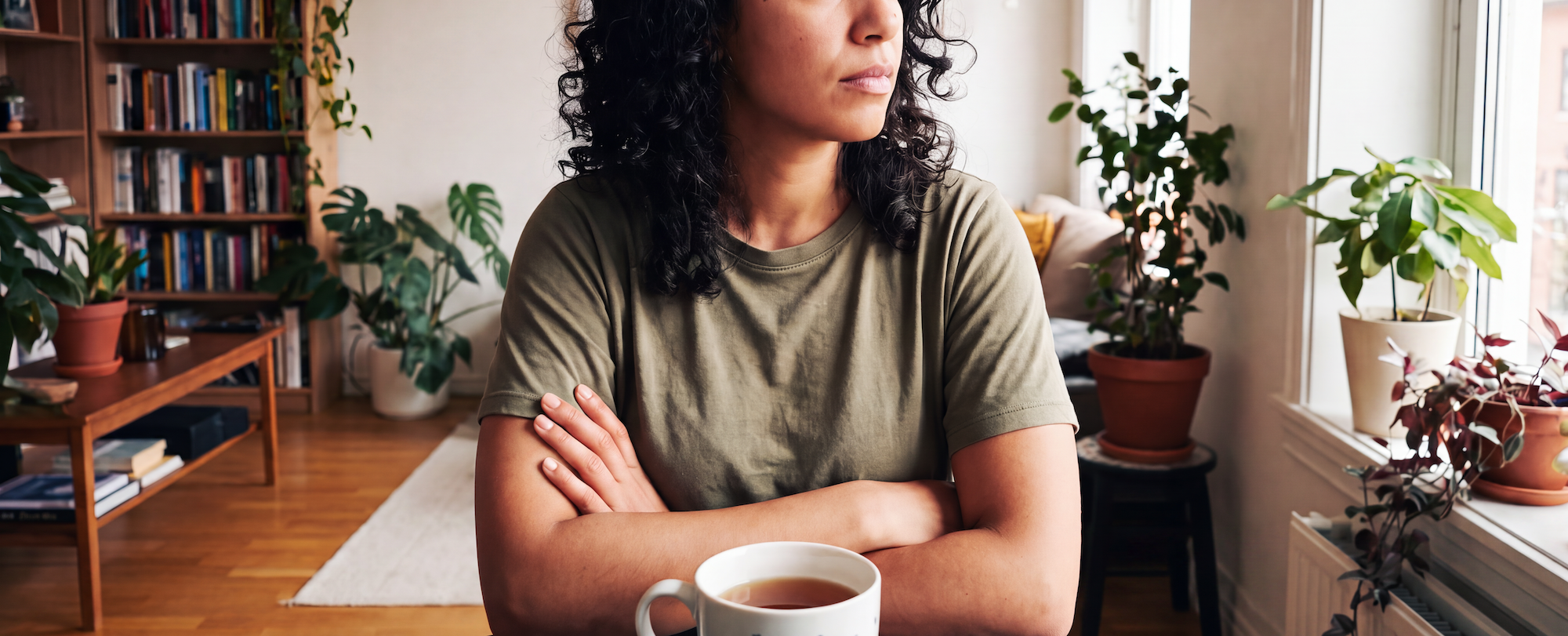 A latina woman in her mid-30's wearing a plain army green tee, sitting at a table with a cup of coffee or tea, with her arms crossed, looking at the window in a contemplative mood; exuding a protecting my peace vibe.